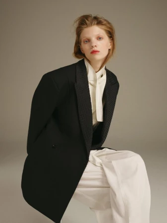 A blond-haired woman in a black blazer and white pants poses in a fashion shot against a neutral grey background.