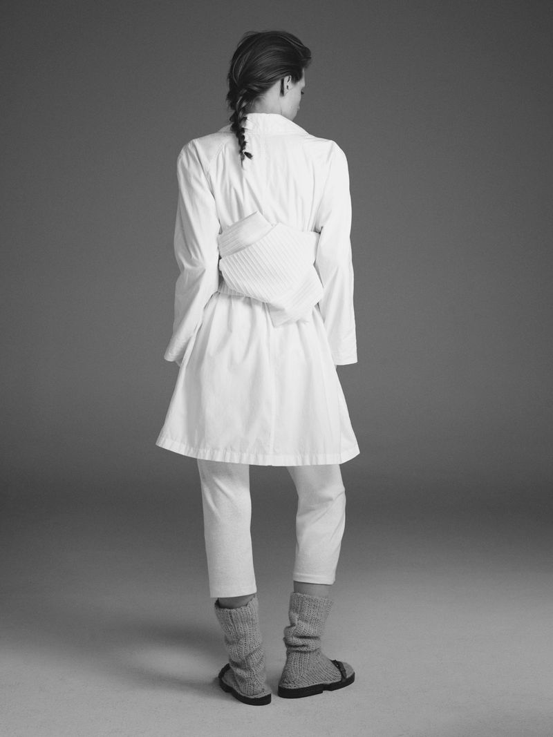 Back view of a woman wearing a white dress and pants with grey knit leg warmers in a studio with a grey background. 