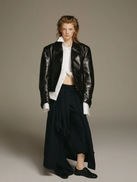 Fashionable young adult female posing in black leather jacket, unbuttoned white shirt, and black skirt in a studio setting.