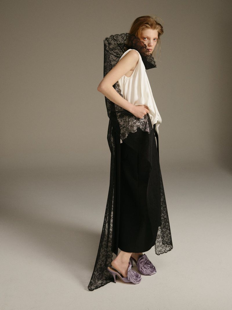 A fashion model poses indoors wearing a white tank top and black lace skirt with matching heels on a neutral grey background.