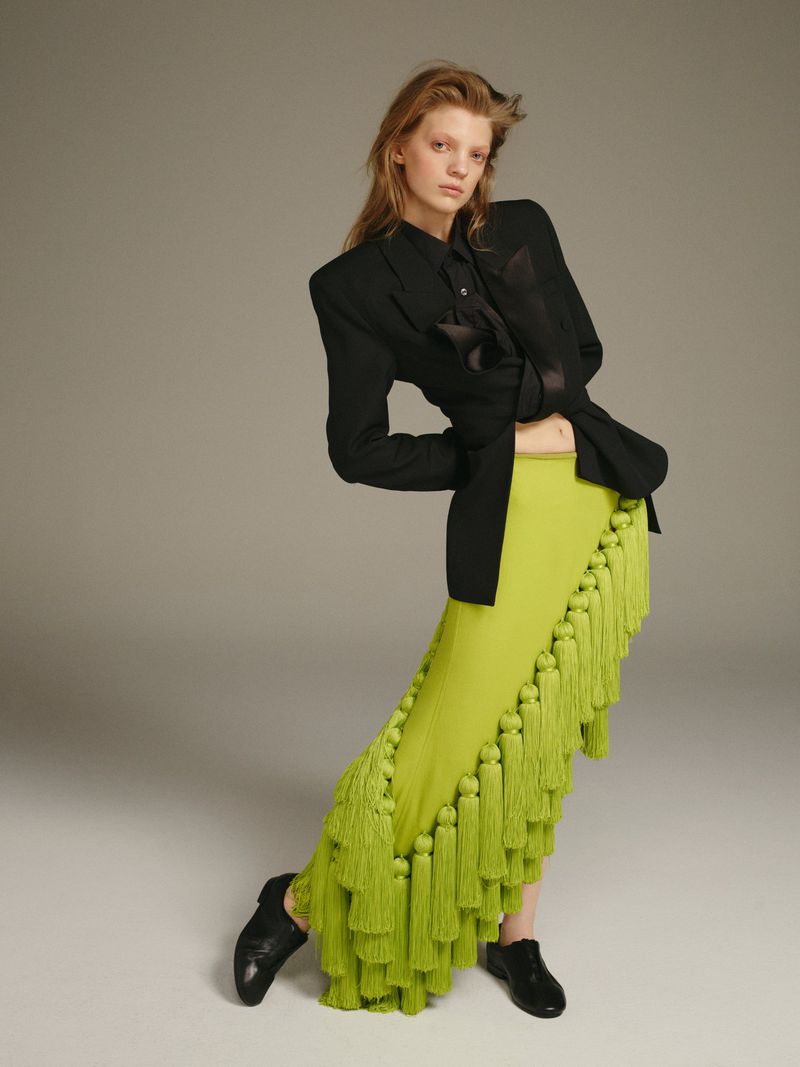 Fashion editorial shot of a young adult woman in a black blazer, button-down shirt, and a long green fringed skirt posing in a studio.