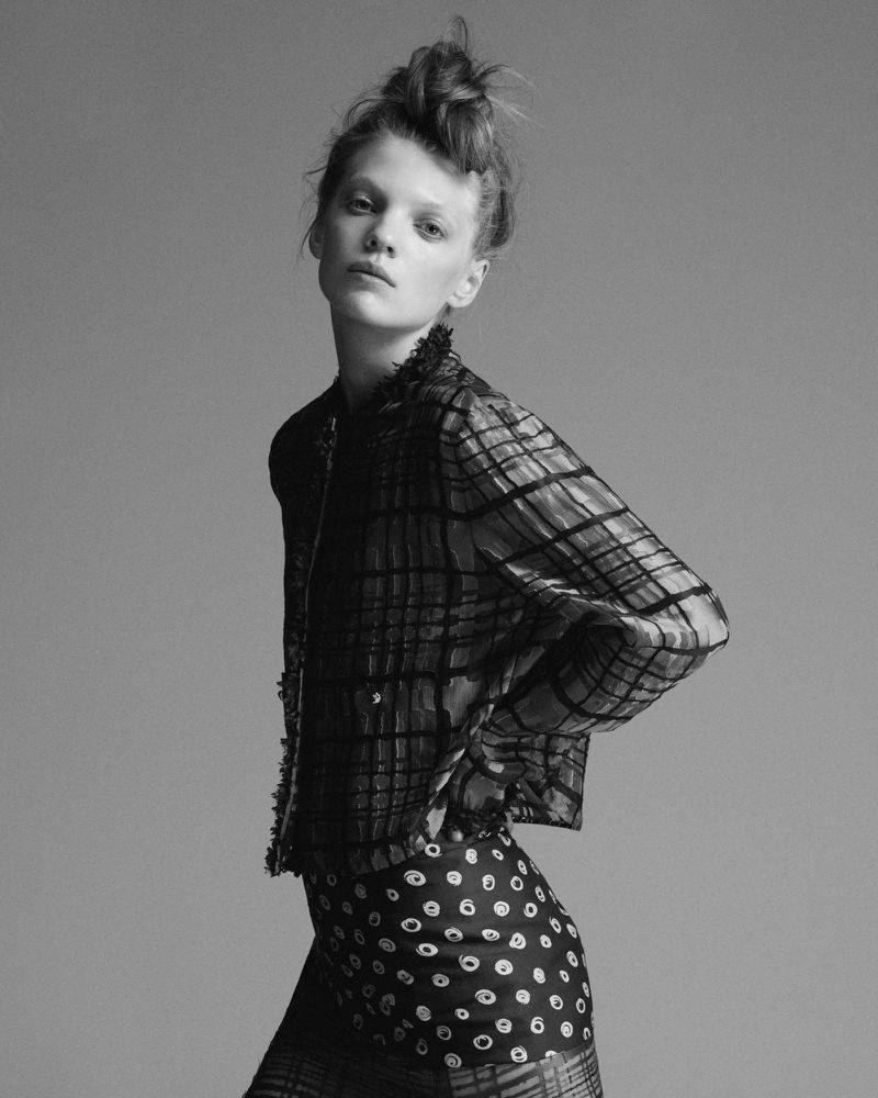 Monochrome portrait of a slender woman with a unique updo, wearing a patterned jacket and skirt, against a plain gray backdrop.