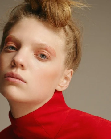 Close-up portrait of a woman with red eye makeup and blonde hair in a bun, wearing a red turtleneck against a neutral background.