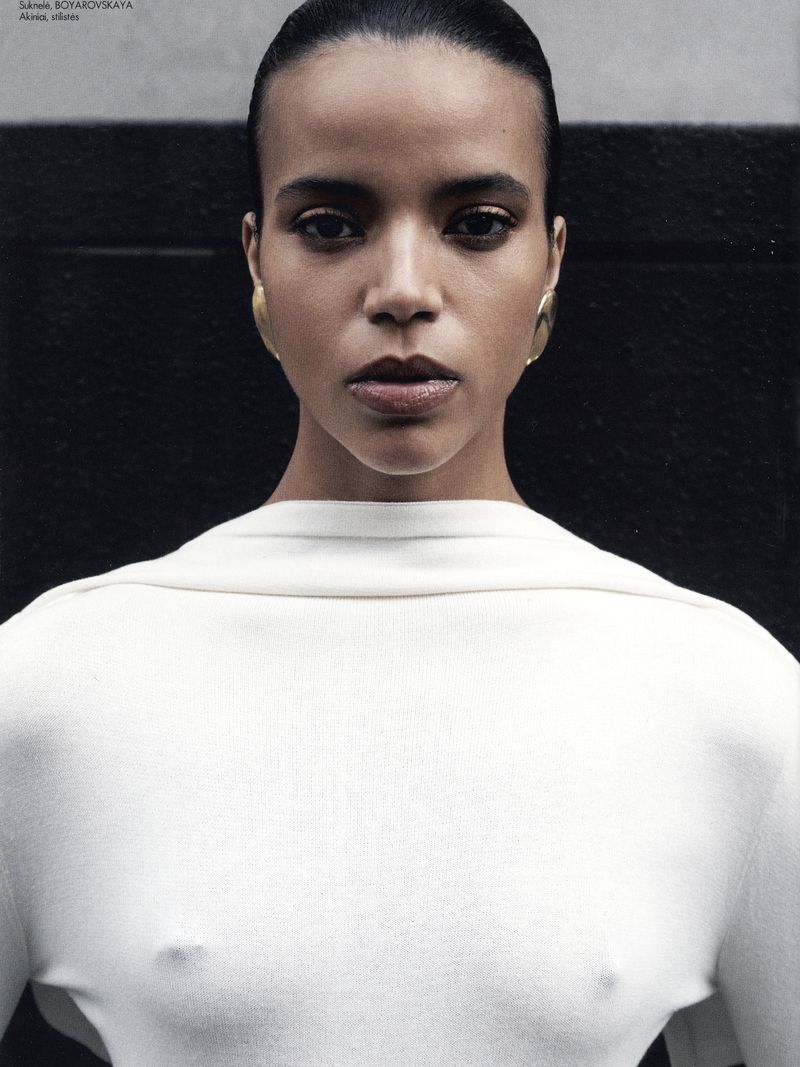 Close-up studio portrait of a woman with slicked-back hair wearing a white long-sleeve shirt and gold hoop earrings against a dark background.