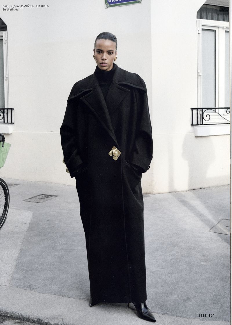 An adult female model in a full-length black coat, black turtleneck, and black heeled boots poses on a city street in a fashion editorial.