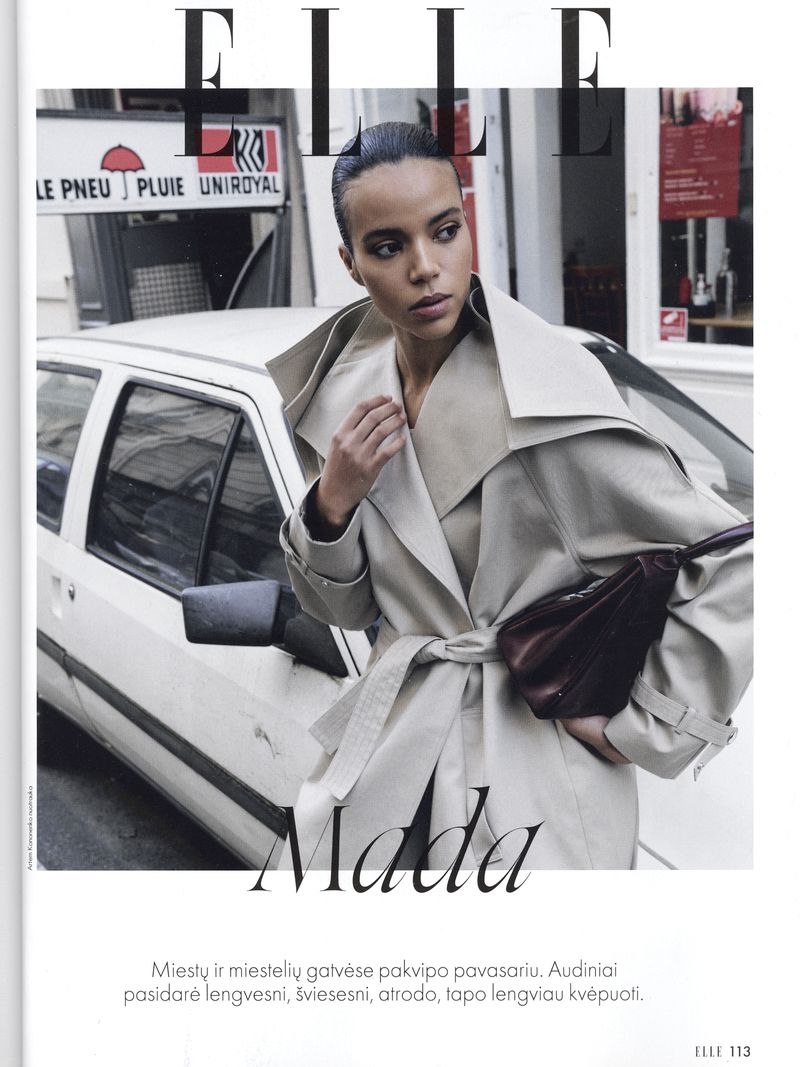 Editorial fashion shot of a woman in a trench coat with a purse, posing near a white car and street signage for a magazine cover.