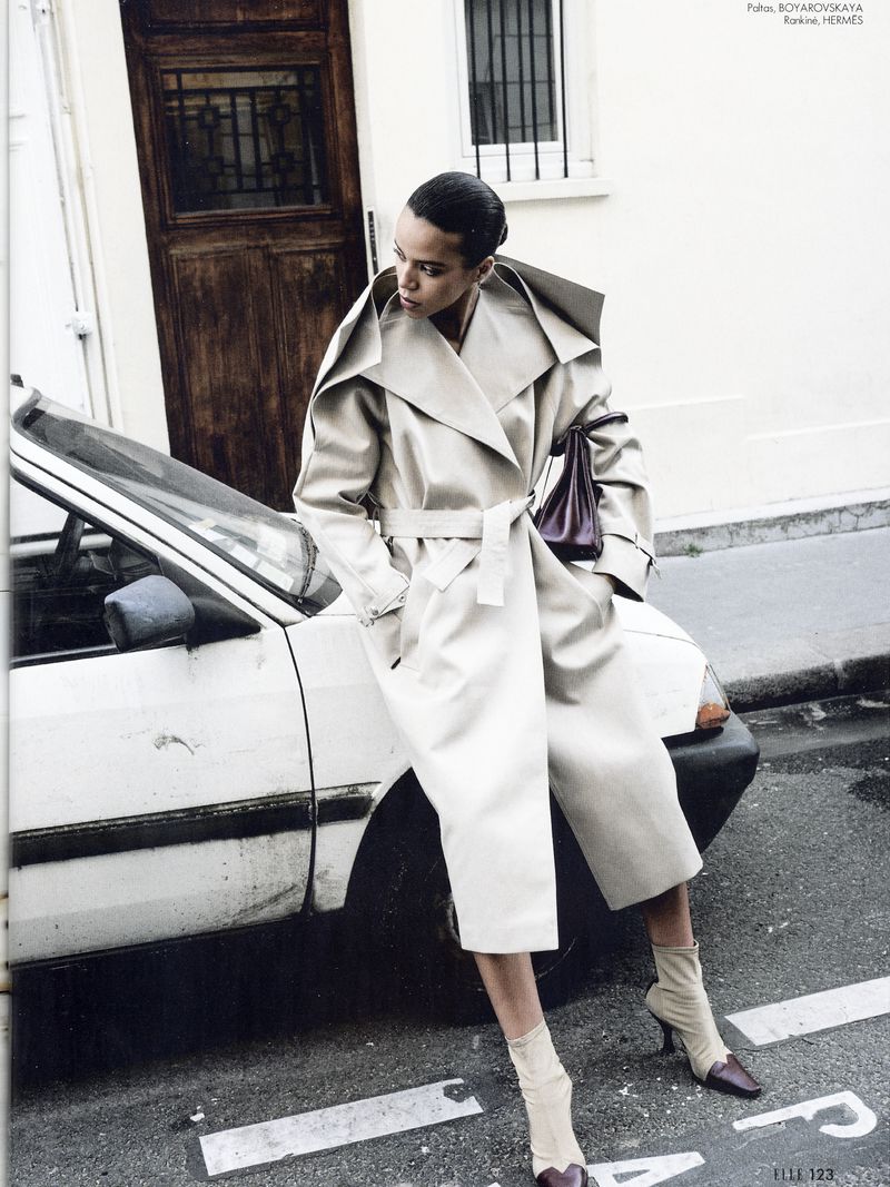 Fashion editorial shot of a woman in a trench coat, bag, and booties leaning against a car near a building in a street setting.