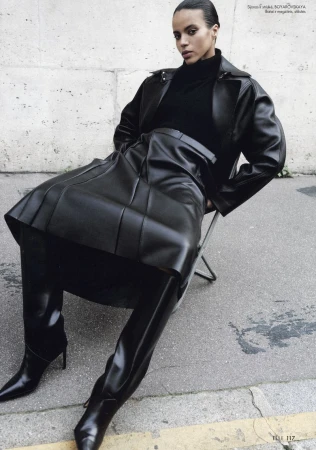 A brunette fashion model in a black leather jacket and skirt sits on a metal folding chair on a street in a fashion editorial.