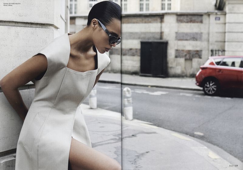 A fashionable woman in an elegant dress and sunglasses poses on a city street with a red car in the background for a fashion editorial.