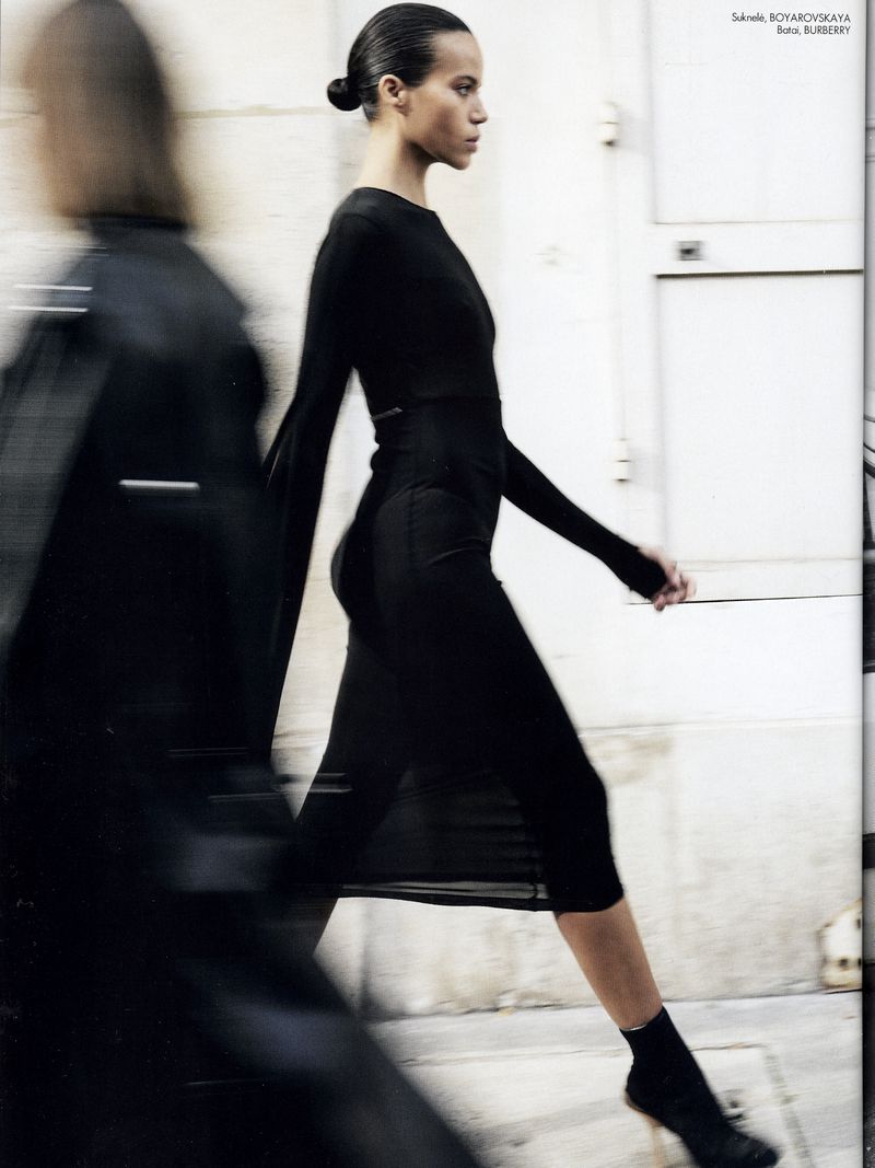 Fashion editorial shot of a woman walking in a long-sleeve black dress and sock boots near a bright grey wall with blurred motion.