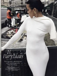 Model in a long white dress with gold earrings posing outdoors on a city street near a construction zone for Elle Lithuania.
