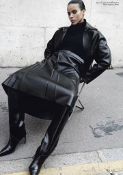 Model wearing a black leather trench coat and skirt seated on a folding chair on a concrete sidewalk.