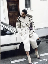 Model leaning against a white vintage car on a city street wearing a structured beige belted trench coat and boots.