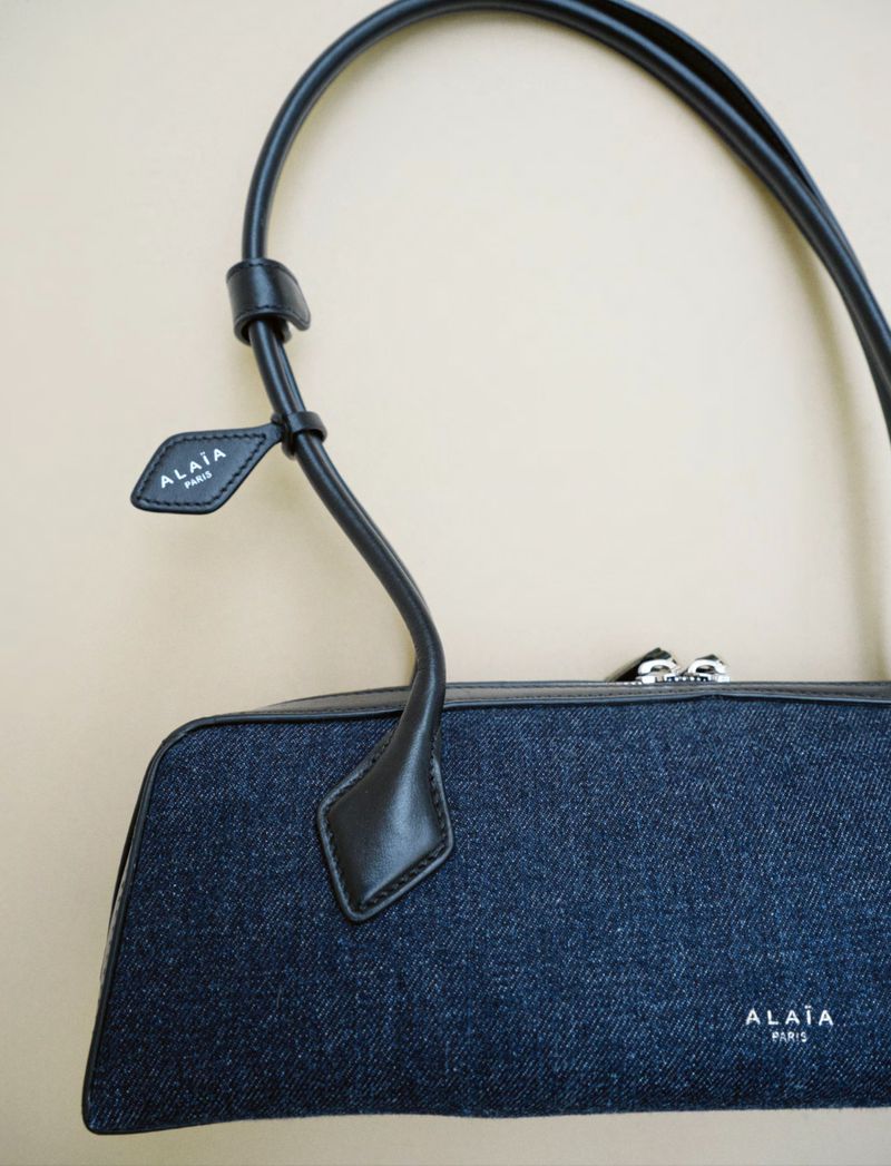 Close-up of a blue denim Alaïa handbag with black leather trim and handles against a neutral background; designer fashion accessory.