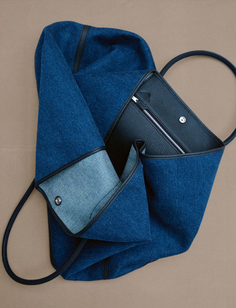 Denim and black leather tote bag with a metal snap closure on a tan background. The bag has a unique design and a casual style.