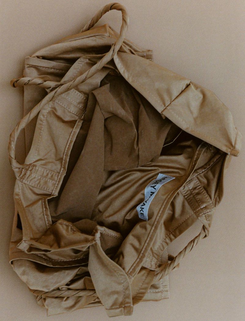 Tan fabric tote bag featuring rope handles and a visible brand label, presented on a neutral background, showcasing its design and material.