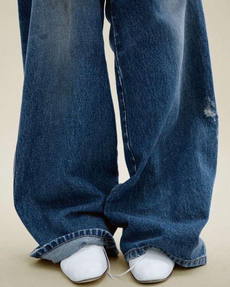 Close-up of distressed, wide-leg denim jeans on a person standing against a neutral background, paired with white, square-toe shoes.