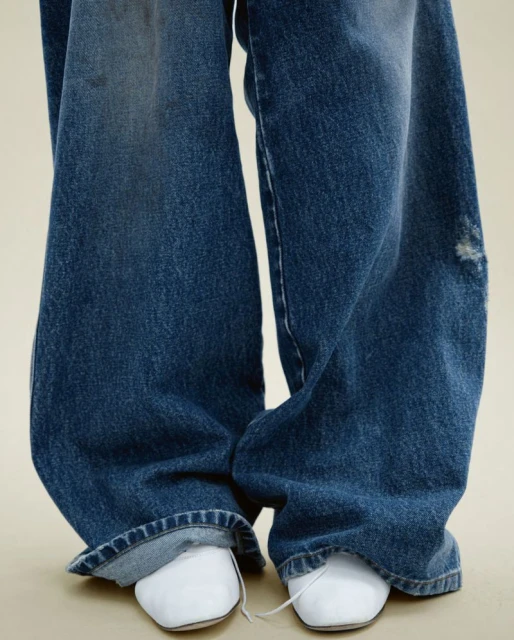 Close-up of distressed, wide-leg denim jeans on a person standing against a neutral background, paired with white, square-toe shoes.