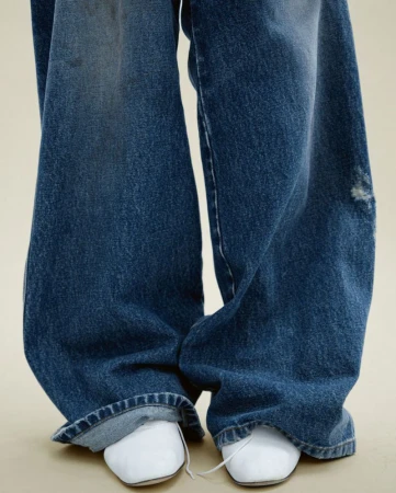 Close-up of distressed, wide-leg denim jeans on a person standing against a neutral background, paired with white, square-toe shoes.