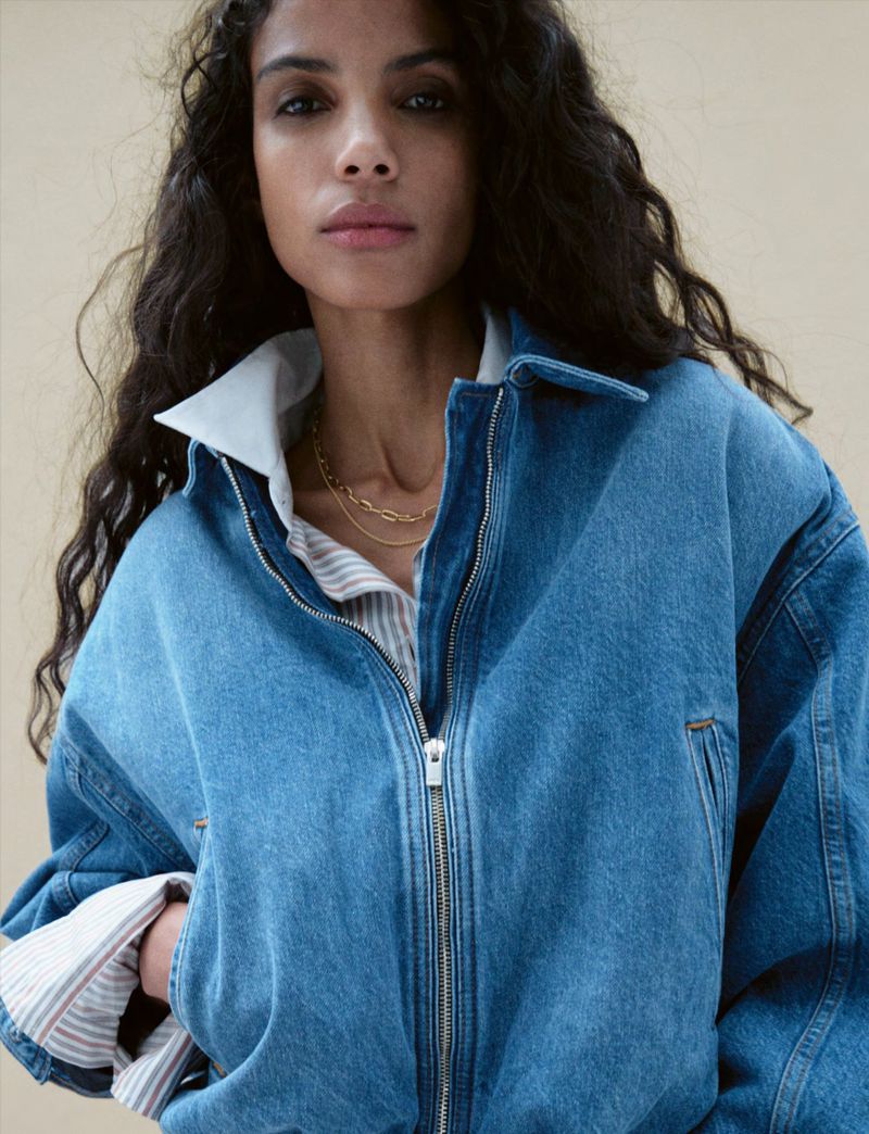 A woman with long wavy dark hair wears a blue denim jacket over a striped shirt and gold chain necklace, looking at the camera.