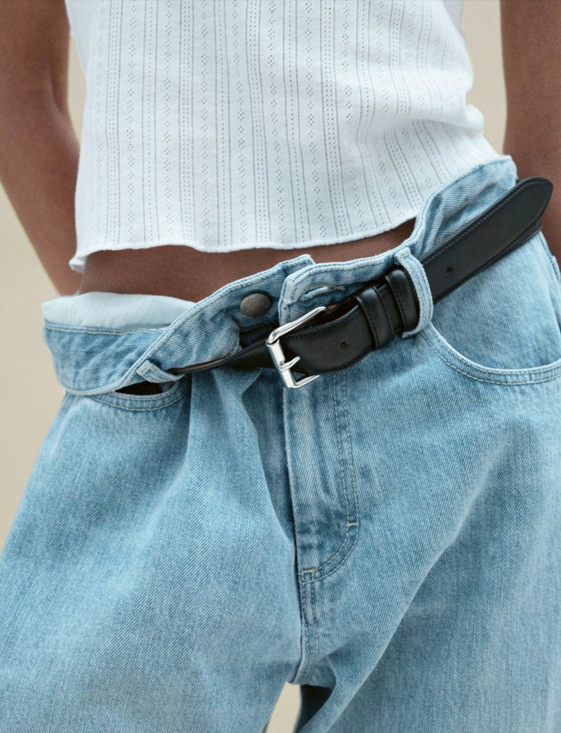 Light blue denim jeans with a black leather belt are styled with a white crop top in a fashion shot. Indoor studio setting.