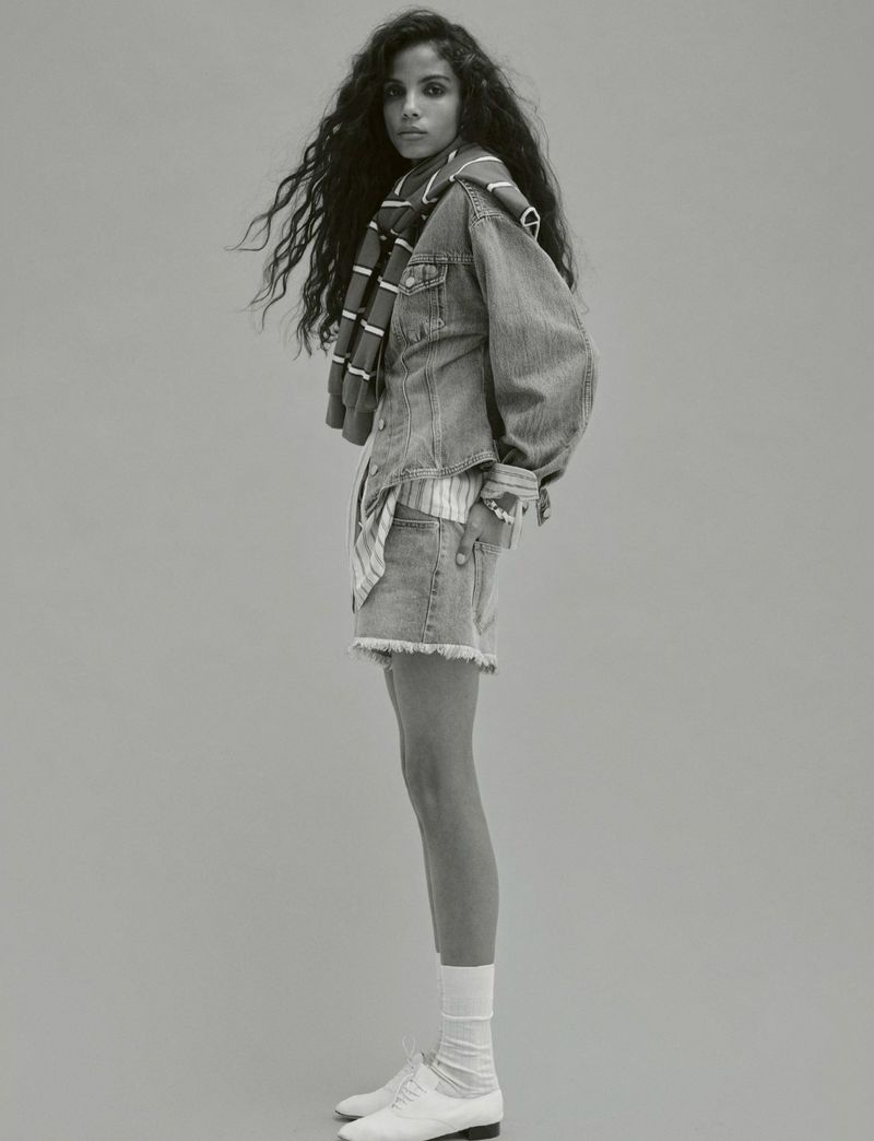 Portrait of a young adult woman with long curly hair wearing a denim outfit with a striped scarf, white socks, and leather shoes.