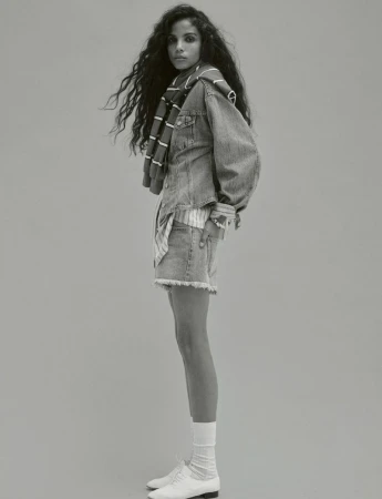 Portrait of a young adult woman with long curly hair wearing a denim outfit with a striped scarf, white socks, and leather shoes.