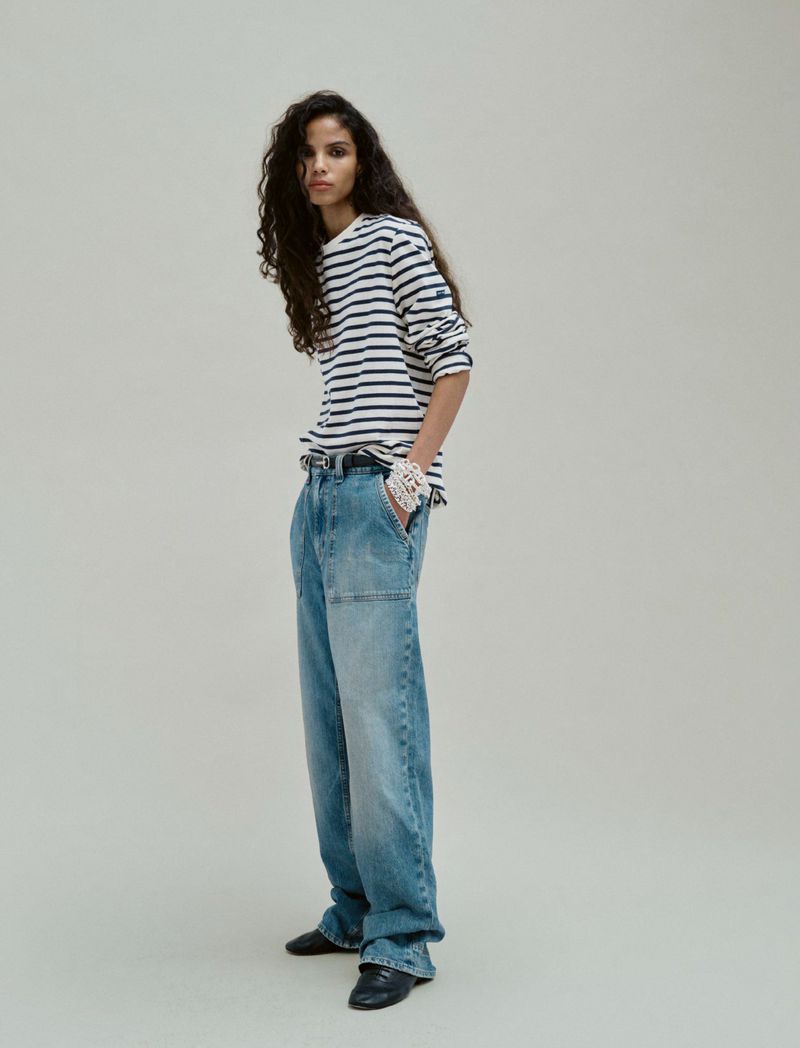 Adult fashion model poses in an indoor photo studio, wearing a striped shirt and loose-fitting denim pants with black flats.