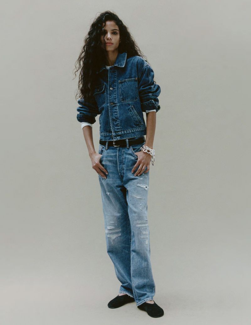 Woman in a denim jacket and distressed jeans poses in a fashion shot. The image has an overall neutral color palette.