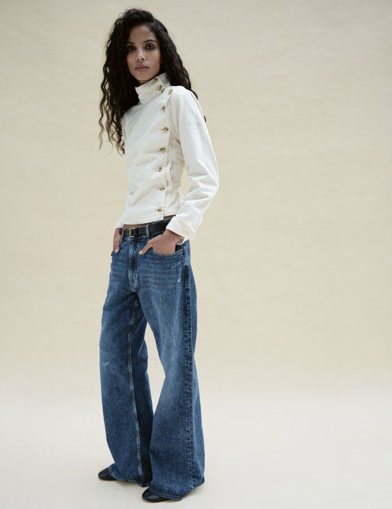 A woman with long, dark curly hair poses in a white, high-necked top with side buttons and wide-leg blue jeans against a beige background.