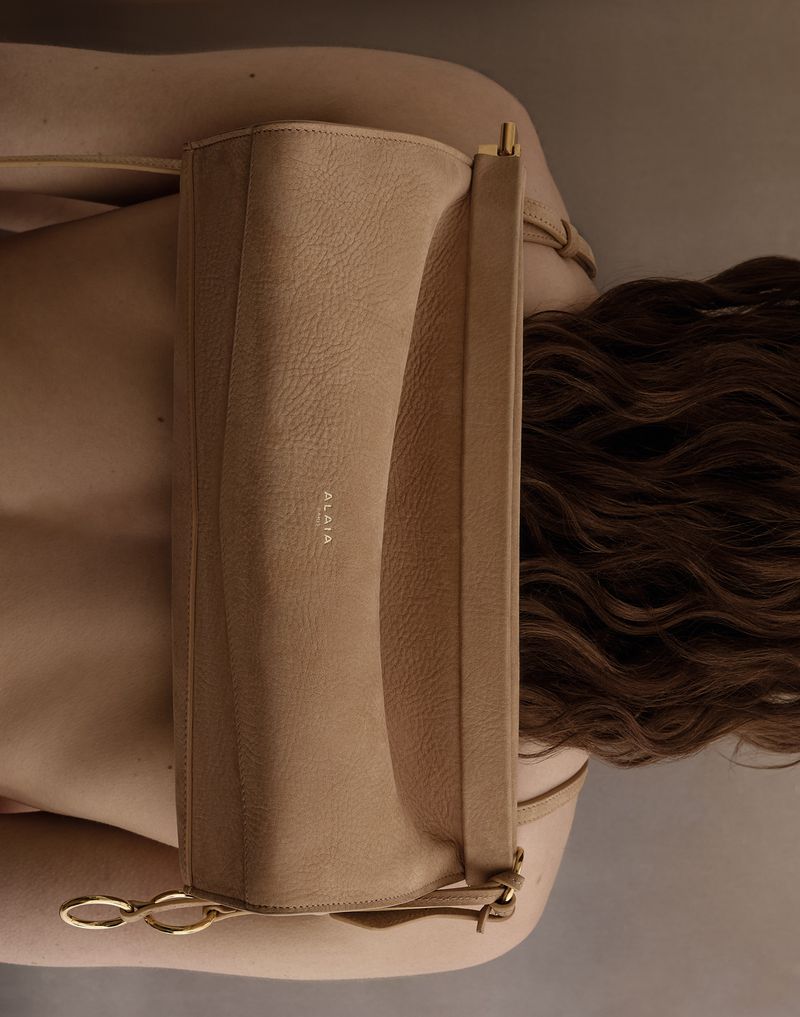 Close-up of a beige Alaia handbag being held against the back of an adult female with wavy brunette hair in a studio setting.