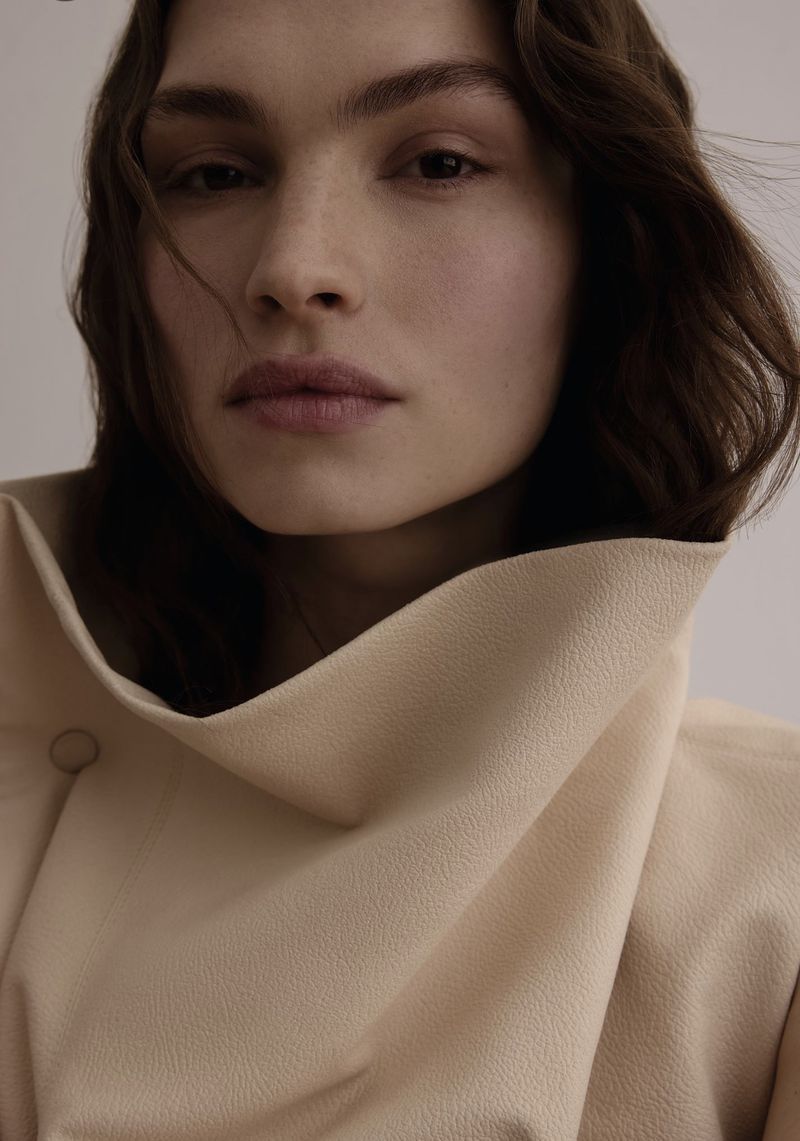 Close-up portrait of an adult woman with wavy brown hair wearing a beige coat with a large collar against a neutral background.