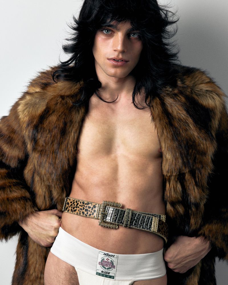Male model with long black hair poses topless in a fur coat and underwear on a white background in a studio fashion shot.