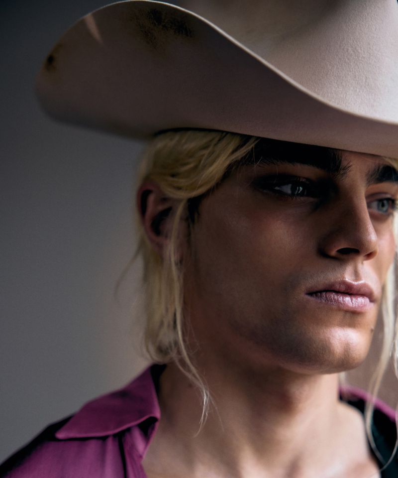 Close-up studio portrait of a young adult male with blond hair wearing a cowboy hat and magenta shirt, looking off to the side.