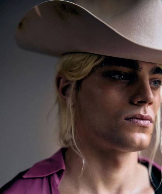 Close-up studio portrait of a young adult male with blond hair wearing a cowboy hat and magenta shirt, looking off to the side.
