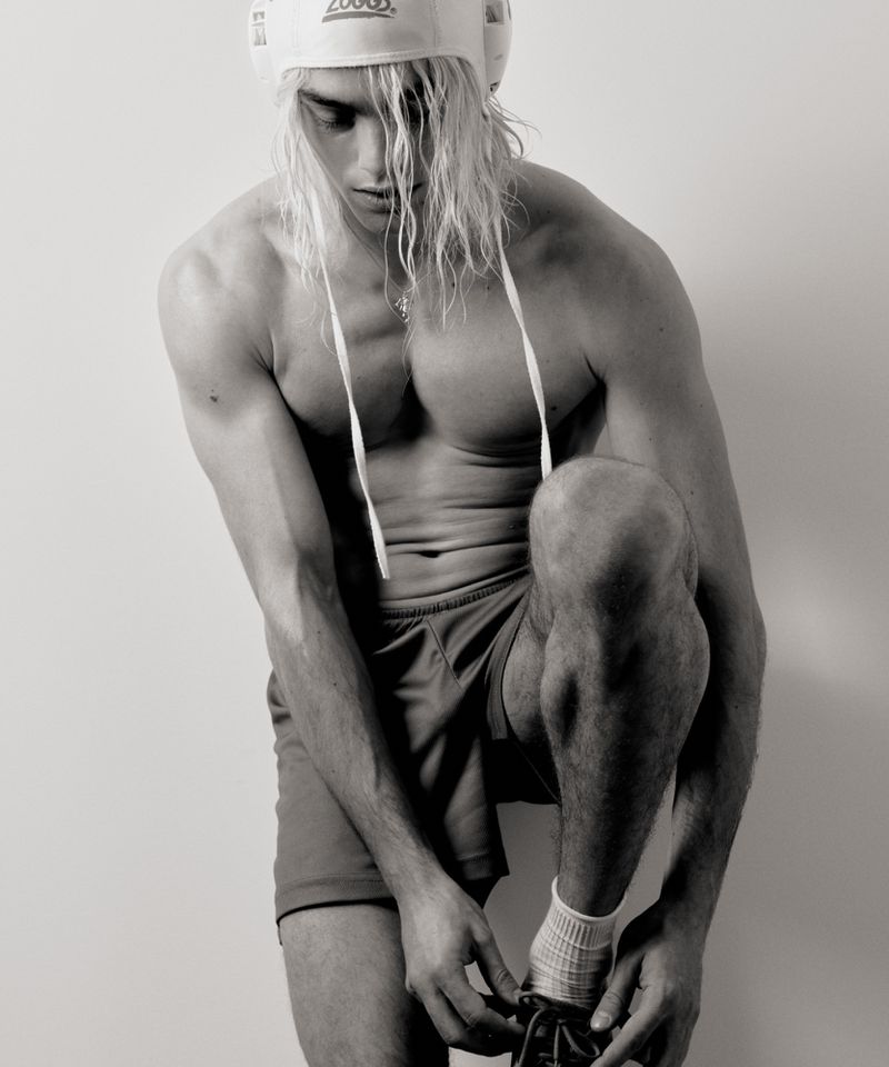 Black and white photo of a fit, shirtless male athlete with long blonde hair and a swim cap, tying the laces of a running shoe.