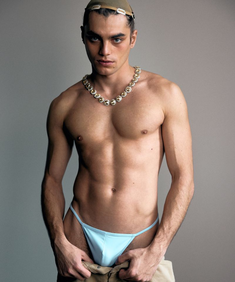 Striking studio portrait of a male model showcasing athletic build with a gold necklace, wearing a light blue underwear, and a tan hat.