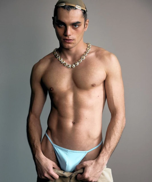 Striking studio portrait of a male model showcasing athletic build with a gold necklace, wearing a light blue underwear, and a tan hat.