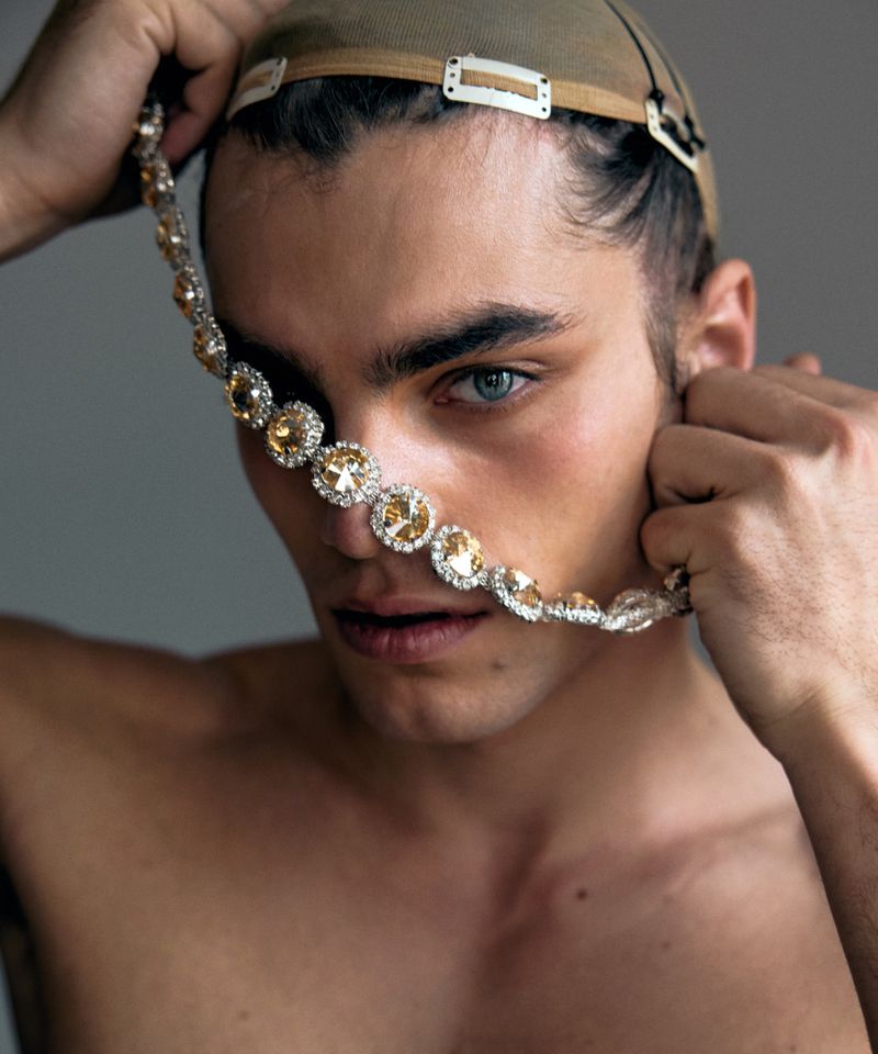Close-up of a young adult male with blue eyes wearing a nude cap and a jeweled necklace covering his mouth, set against a grey backdrop.