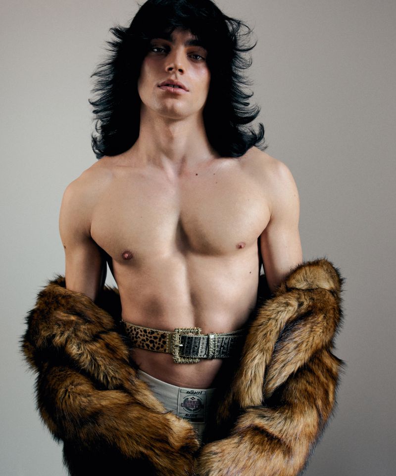 Shirtless man with long black hair and a fur coat poses indoors with a patterned belt. Fashion and portrait photography.