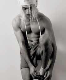 Shirtless male model wearing swim cap and shorts tying shoe against a neutral studio background