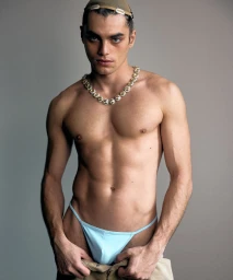 Shirtless male model wearing a light blue thong and a chunky gemstone necklace against a neutral grey studio background.