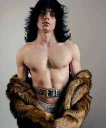 Shirtless male model with long dark hair wearing a patterned belt and fur jacket against a plain grey background