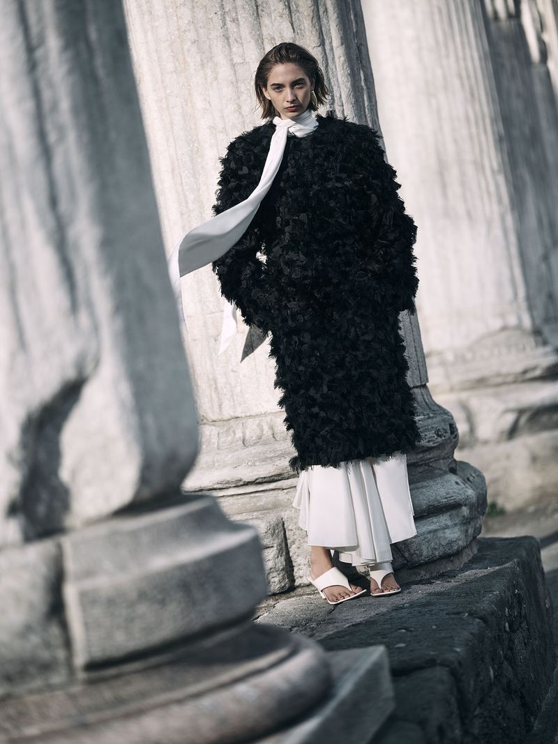 A woman in a black textured coat and white pants stands on stone steps near columns in natural light, in a fashion editorial.
