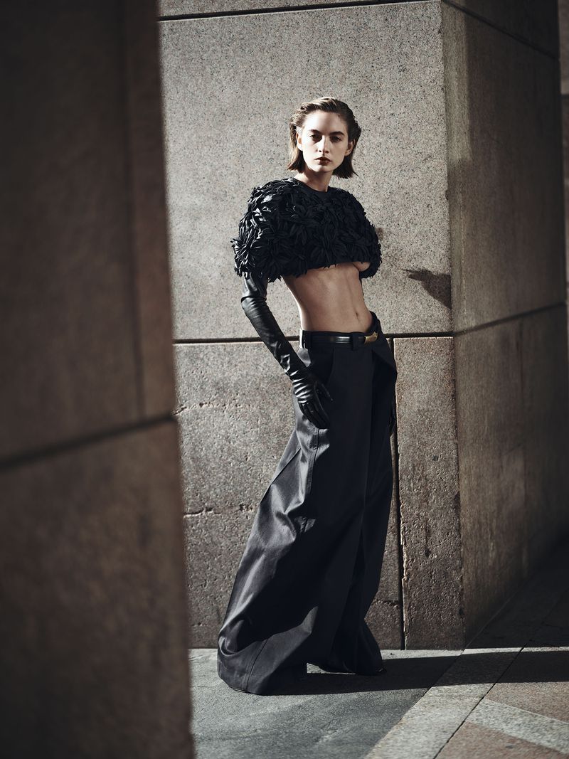 Model wearing a black textured cropped top and long trousers standing against a stone pillar background