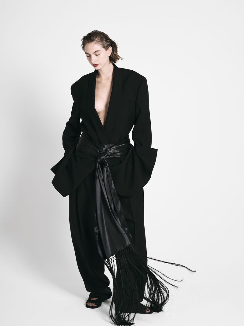 A woman with wet hair wearing a black oversized blazer and matching trousers with a long satin fringed sash against a white background.