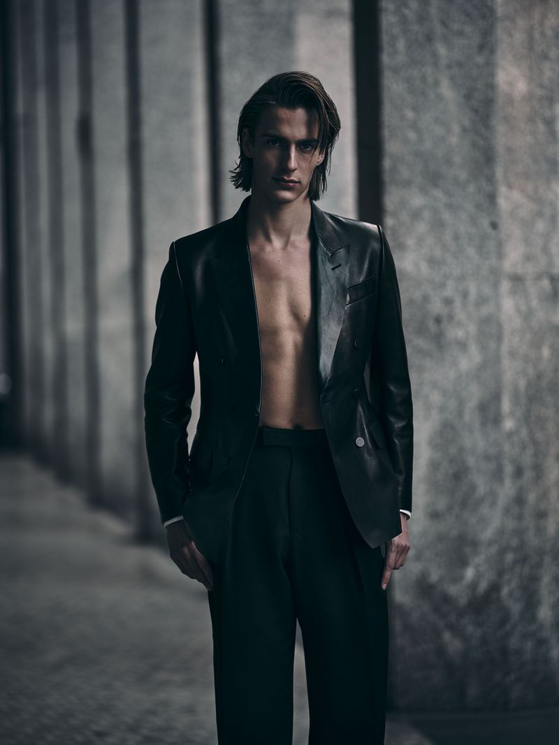 Portrait of a male model in a dark suit with open jacket posing against a concrete wall in dim lighting indoors.