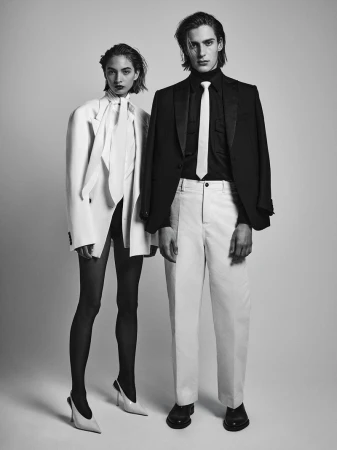 Black and white editorial fashion portrait of a female model in a white blazer and a male model in a black suit with a white tie.