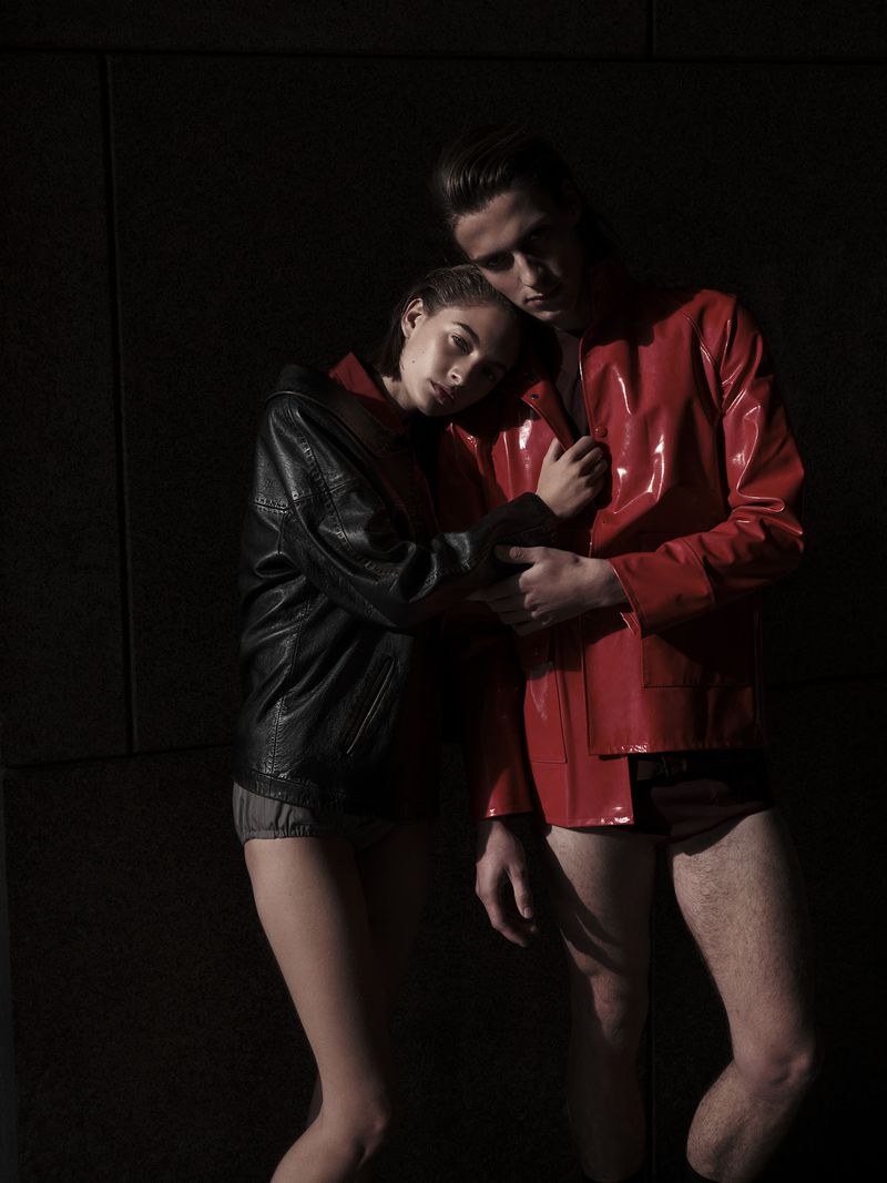 A man and woman, wearing matching shiny red and black jackets and dark bottoms, pose intimately against a dark, textured wall.