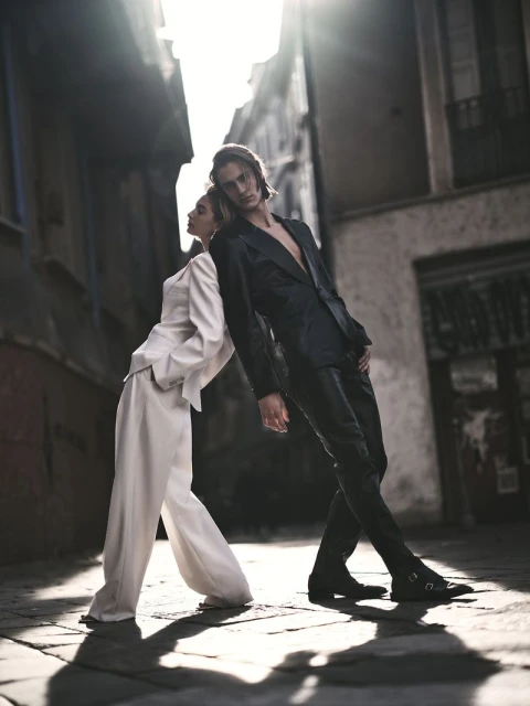 Fashionable couple in suit leans back to back in sunlit alleyway. Woman in cream suit, man in black suit with chest exposed.